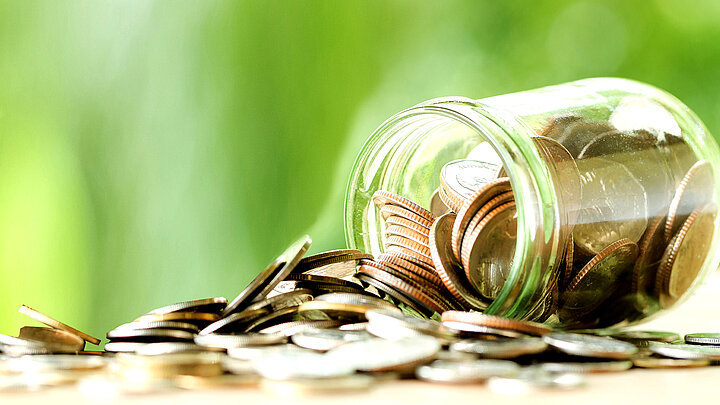 Grafika dekoracyjna. Przewrócony słoik, z którego wysypują się monety i pokrywają całą dolna część obrazka. Z tyłu zielone tło.