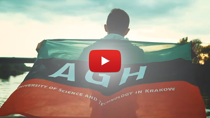 Zdjęcie przedstawia młodego mężczyznę zwróconego tyłem do obserwatora. Mężczyzna trzyma powiewającą na wietrze trójkolorową flagę z literami AGH.